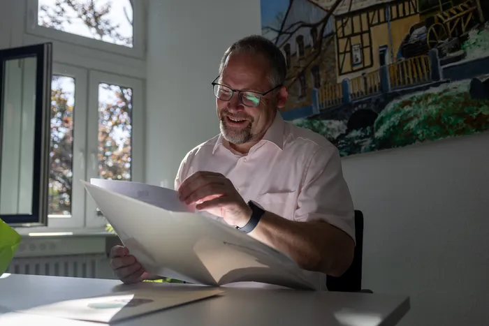 Ein Mann mit Brille und Hemd liest ein geöffnetes Dokument an einem Schreibtisch. Im Hintergrund ein Bild von einem Fachwerkhause.