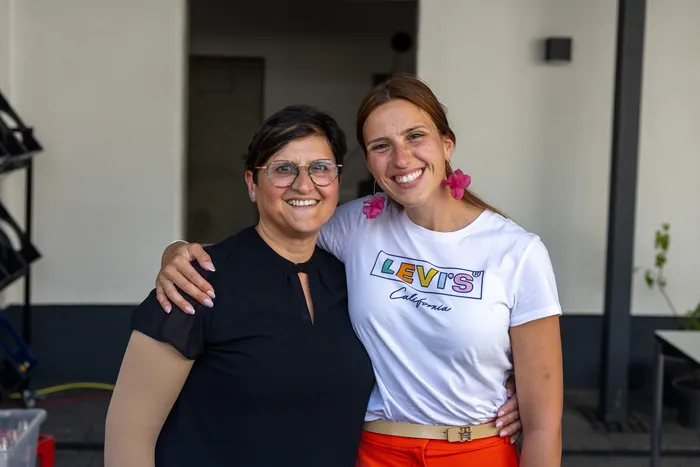 Zwei lächelnde Frauen umarmen sich vor einem hellen Gebäude. Die jüngere Frau trägt ein weißes T-Shirt mit dem Levi's-Logo und orangefarbene Hosen.