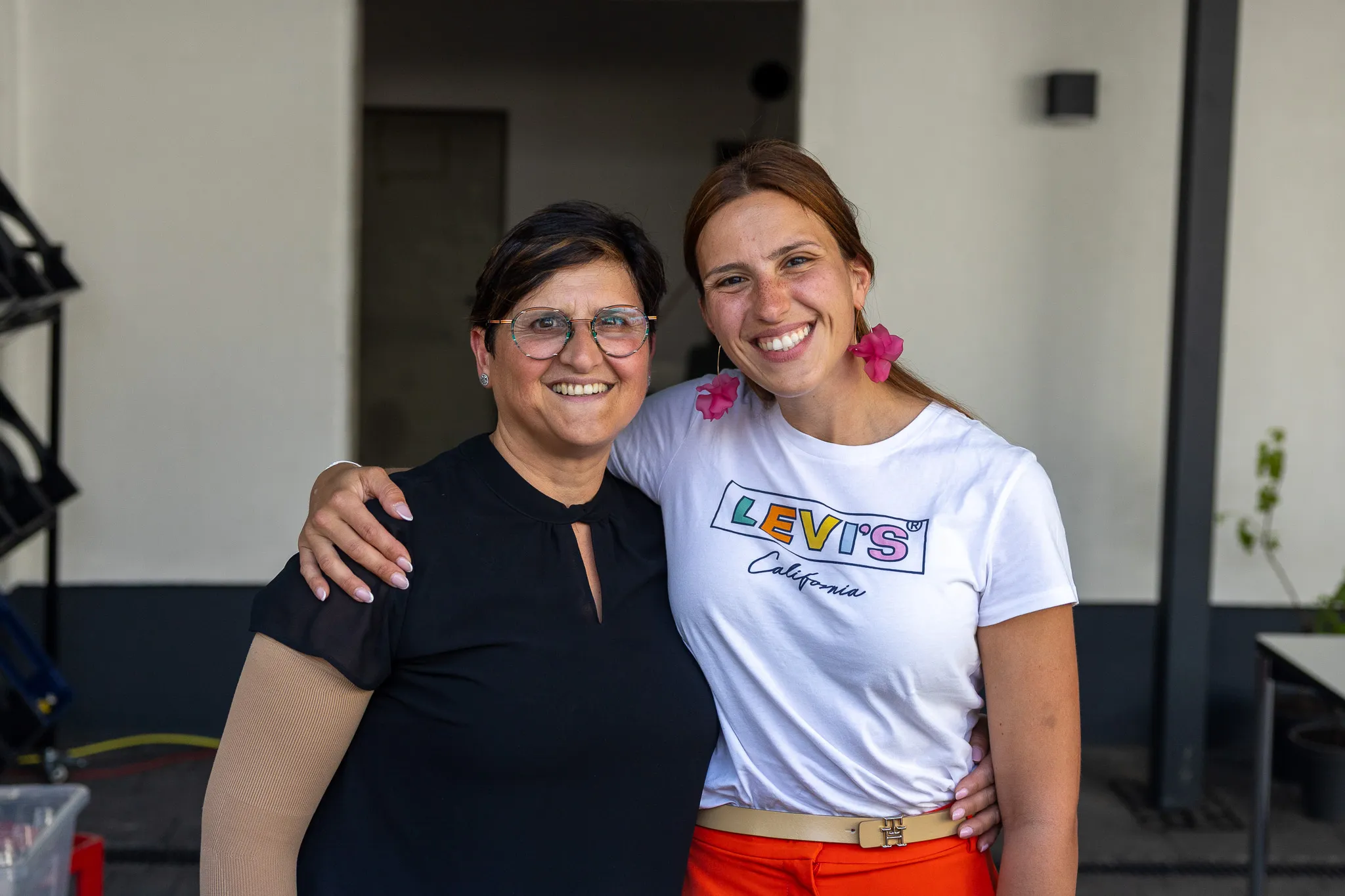Zwei lächelnde Frauen umarmen sich vor einem hellen Gebäude. Die jüngere Frau trägt ein weißes T-Shirt mit dem Levi's-Logo und orangefarbene Hosen.