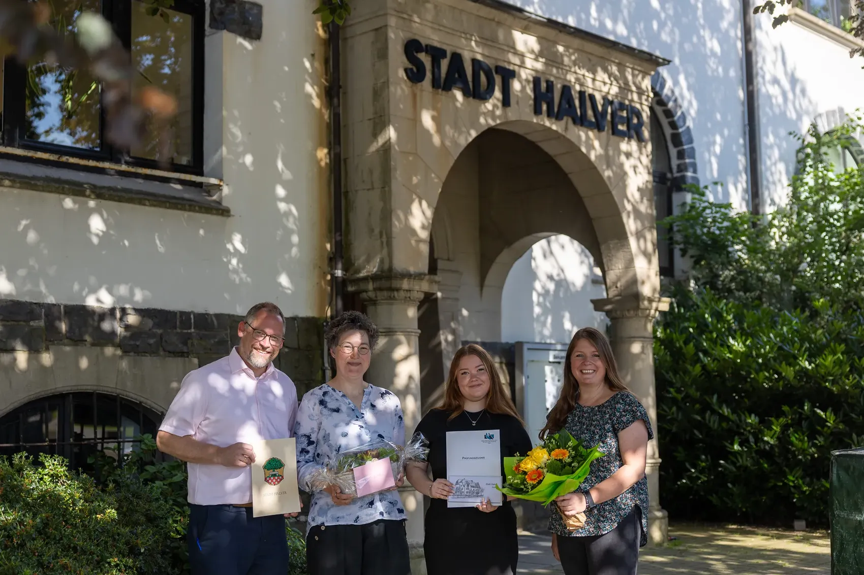 Vier Personen stehen vor dem Haupteingang des Halverer Rathauses. Von links: Bürgermeister Armin Kibbert, Fachbereichsleiterin Annette Schulte, die frischgebackene Verwaltungsfachangestellte Jolina Klotz und Ausbildungsleiterin Elke Gierse. Jolina Klotz und Annette Schulte halten Blumensträuße und Urkunden.