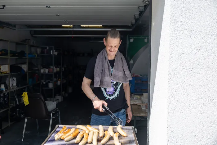 Ein Mann grillt Würstchen vor einer offenen Garagentür, während im Hintergrund Regale mit verschiedenen Gegenständen zu sehen sind.