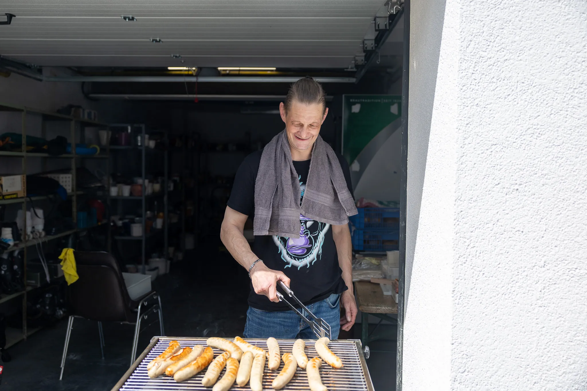 Ein Mann grillt Würstchen vor einer offenen Garagentür, während im Hintergrund Regale mit verschiedenen Gegenständen zu sehen sind.