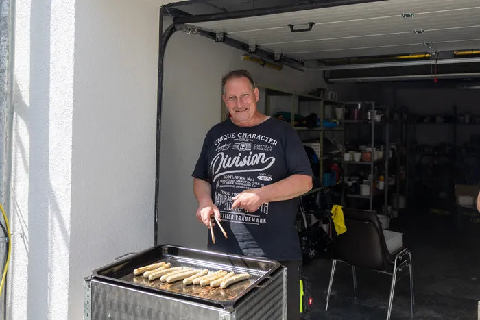 Ein Mann steht lächelnd vor einem Grill mit Bratwürsten und bedient ihn mit einer Zange. Im Hintergrund ist eine offene Garagentür zu sehen.