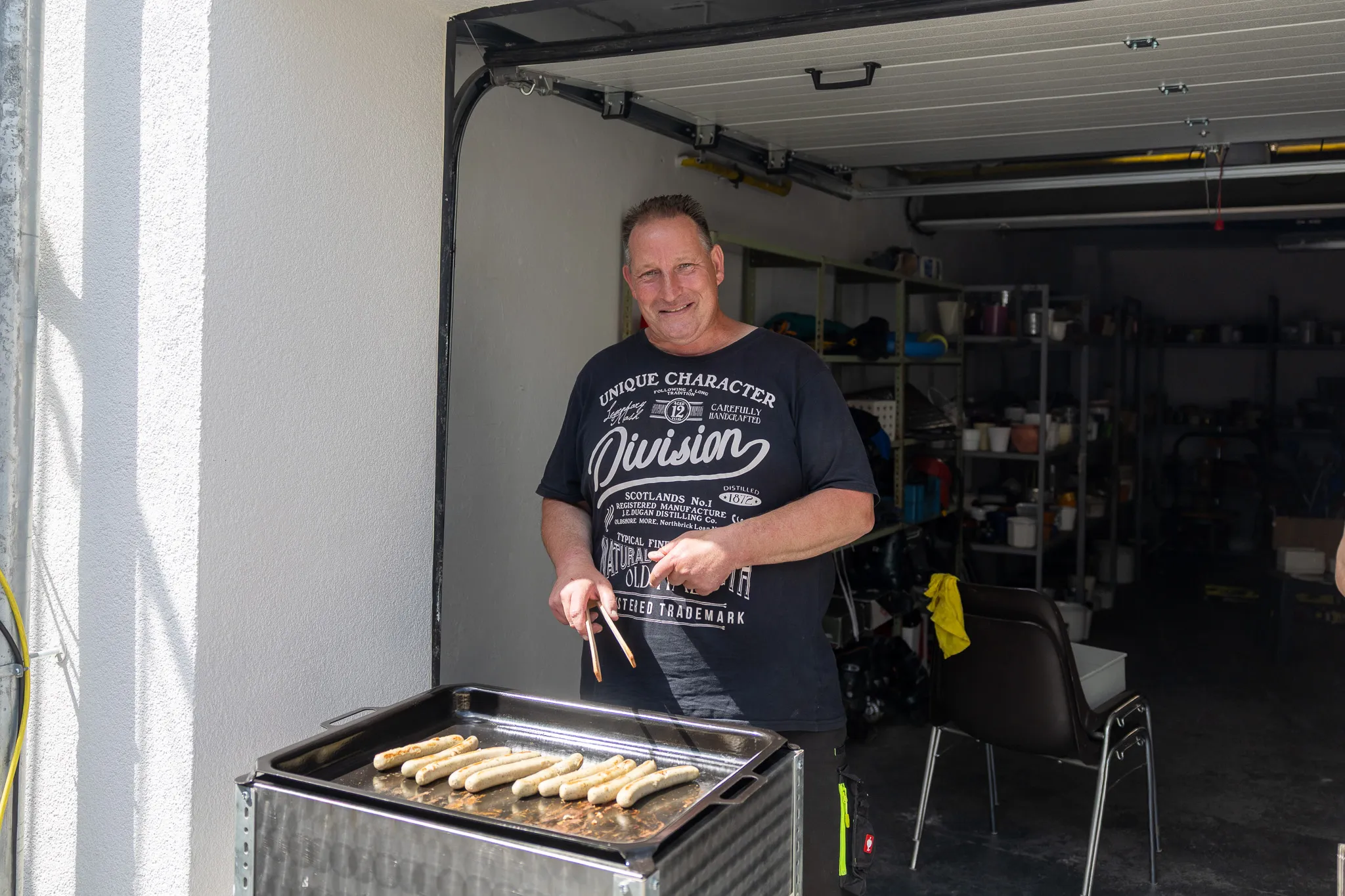 Ein Mann steht lächelnd vor einem Grill mit Bratwürsten und bedient ihn mit einer Zange. Im Hintergrund ist eine offene Garagentür zu sehen.