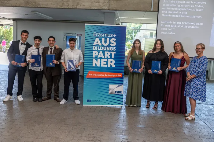 Eine Gruppe junger Erwachsener steht vor einem Banner mit der Aufschrift „Erasmus+ AUS BILDUNGSPARTNER“ und hält jeweils eine blaue Mappe in der Hand.