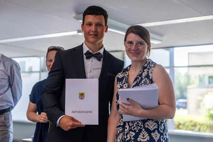Zwei junge Erwachsene, ein Mann in einem dunklen Anzug und eine Frau in einem geblümten Kleid, halten jeweils eine Urkunde mit dem Wort 