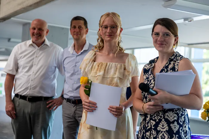 Vier Personen stehen nebeneinander und blicken in die Kamera. Eine junge Frau in einem hellen Kleid hält ein Dokument und eine gelbe Rose. Neben ihr steht eine Frau mit einem Dokument und einem Mikrofon. Im Hintergrund stehen zwei Männer.