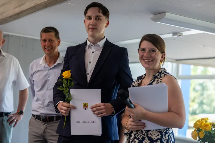 Ein junger Mann in Anzug und mit gelber Rose hält sein Abiturzeugnis. Neben ihm stehen strahlend ein Mann und eine Frau.