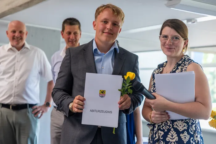 Ein junger Mann in einem grauen Anzug hält ein Abiturzeugnis und einen Blumenstrauß vor einem Mikrofon. Im Hintergrund stehen lächelnde Erwachsene.
