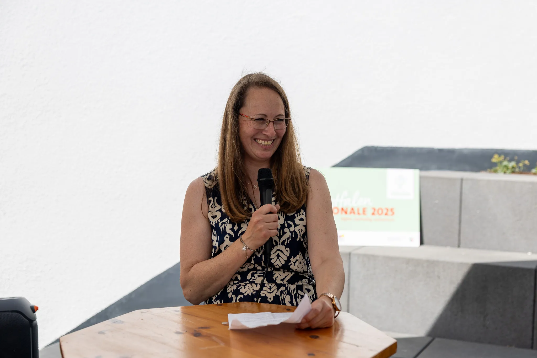 Lachende Frau mit Brille spricht in ein Mikrofon während einer Veranstaltung im Freien. Im Hintergrund ein Banner mit der Aufschrift „Hafen.onale 2025“.