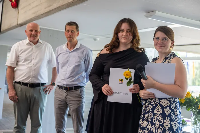 Eine junge Frau hält eine Urkunde mit der Aufschrift „Abiturzeugnis“ und eine gelbe Rose. Neben ihr stehen zwei Lehrer und ein weiterer Mann, vermutlich ebenfalls eine Lehrkraft.
