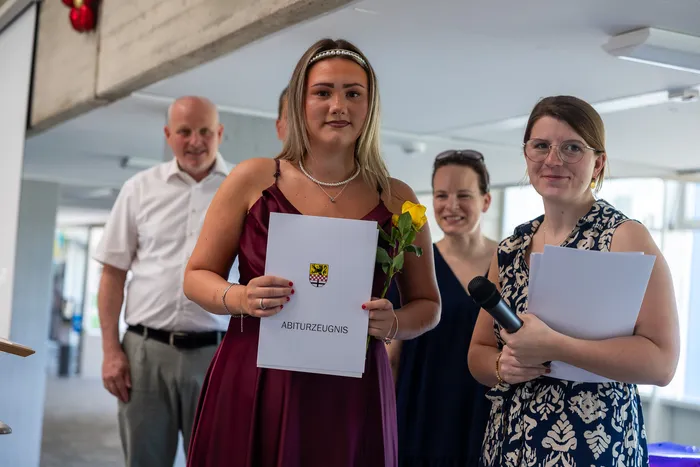 Eine junge Frau in einem roten Kleid hält ihr Abiturzeugnis und eine gelbe Rose. Im Hintergrund stehen ein Mann, eine Frau und eine weitere Person, die in die Kamera blickt.