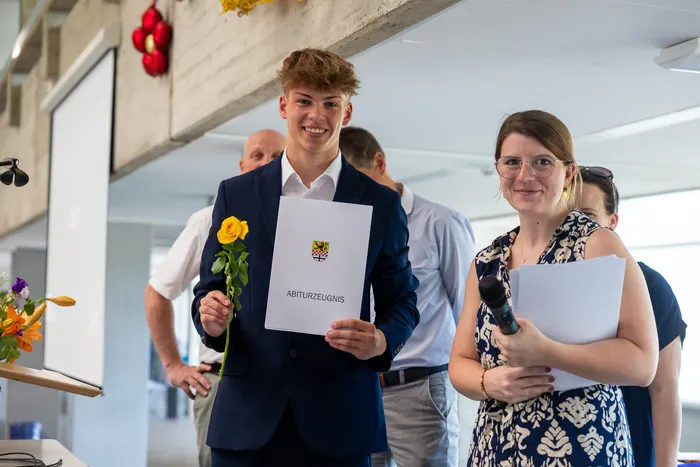 Ein junger Mann in einem dunklen Anzug hält eine gelbe Rose und ein Abiturzeugnis mit dem Wappen des Gymnasiums. Neben ihm steht eine Frau mit einem Mikrofon in der Hand und einem weiteren Abiturzeugnis.