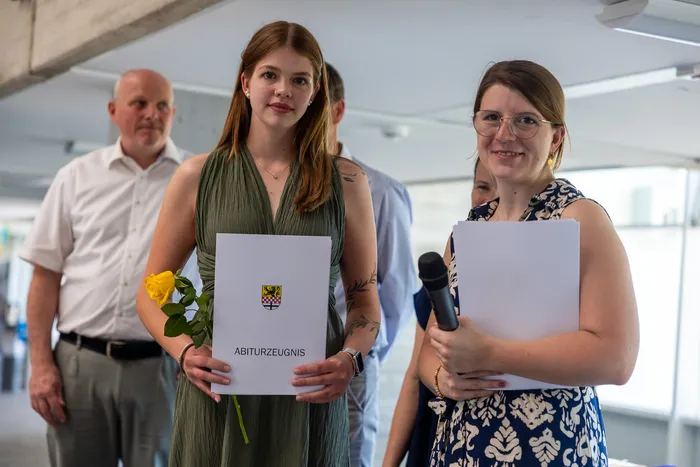 Zwei junge Frauen halten ihre Abiturzeugnisse und eine gelbe Rose. Im Hintergrund steht ein Mann.