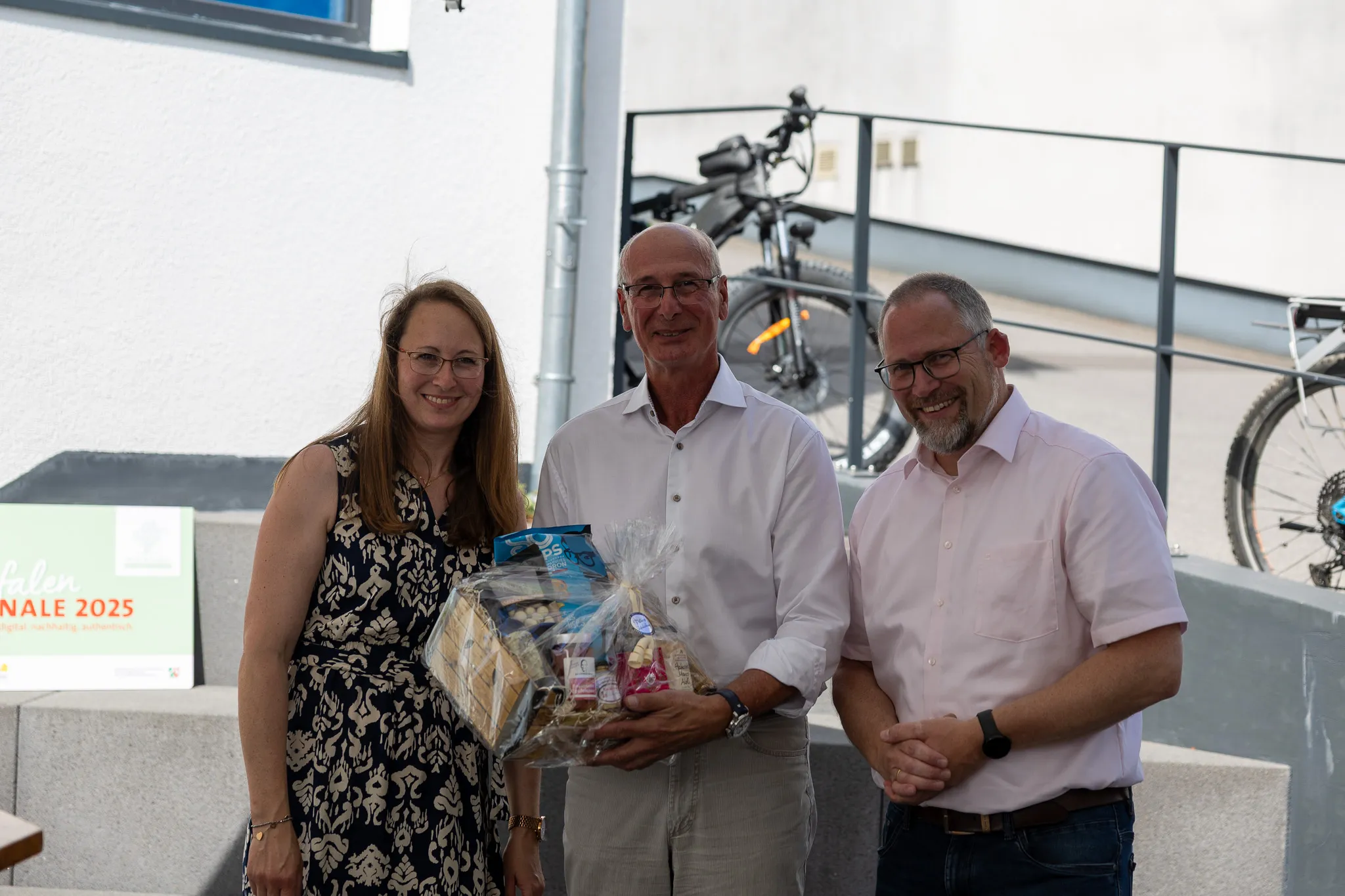 Drei Personen lächeln in die Kamera, der Mann in der Mitte hält einen Korb mit Lebensmitteln und kleinen Geschenken. Im Hintergrund sind Fahrräder zu sehen.