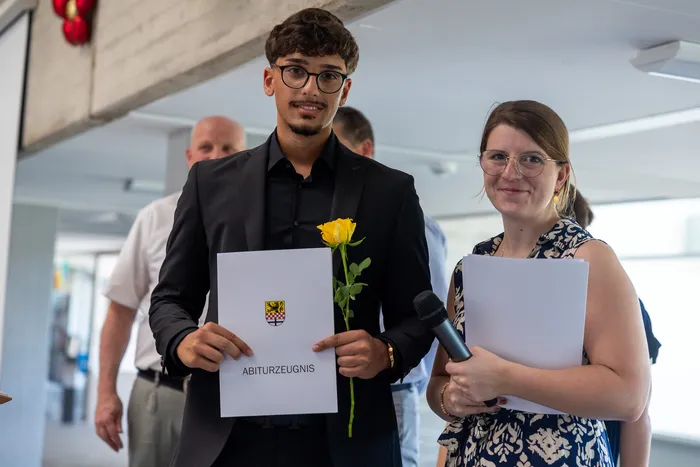 Ein junger Mann mit dunklen Haaren und Brille hält ein Abiturzeugnis und eine gelbe Rose. Neben ihm steht eine junge Frau mit Brille und lächelt in die Kamera.