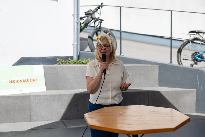 Eine Frau mit blonden Haaren hält ein Mikrofon und spricht auf einer Veranstaltung vor Publikum. Im Hintergrund sind Fahrräder und ein Banner mit der Aufschrift „Regionale 2025“ zu sehen.