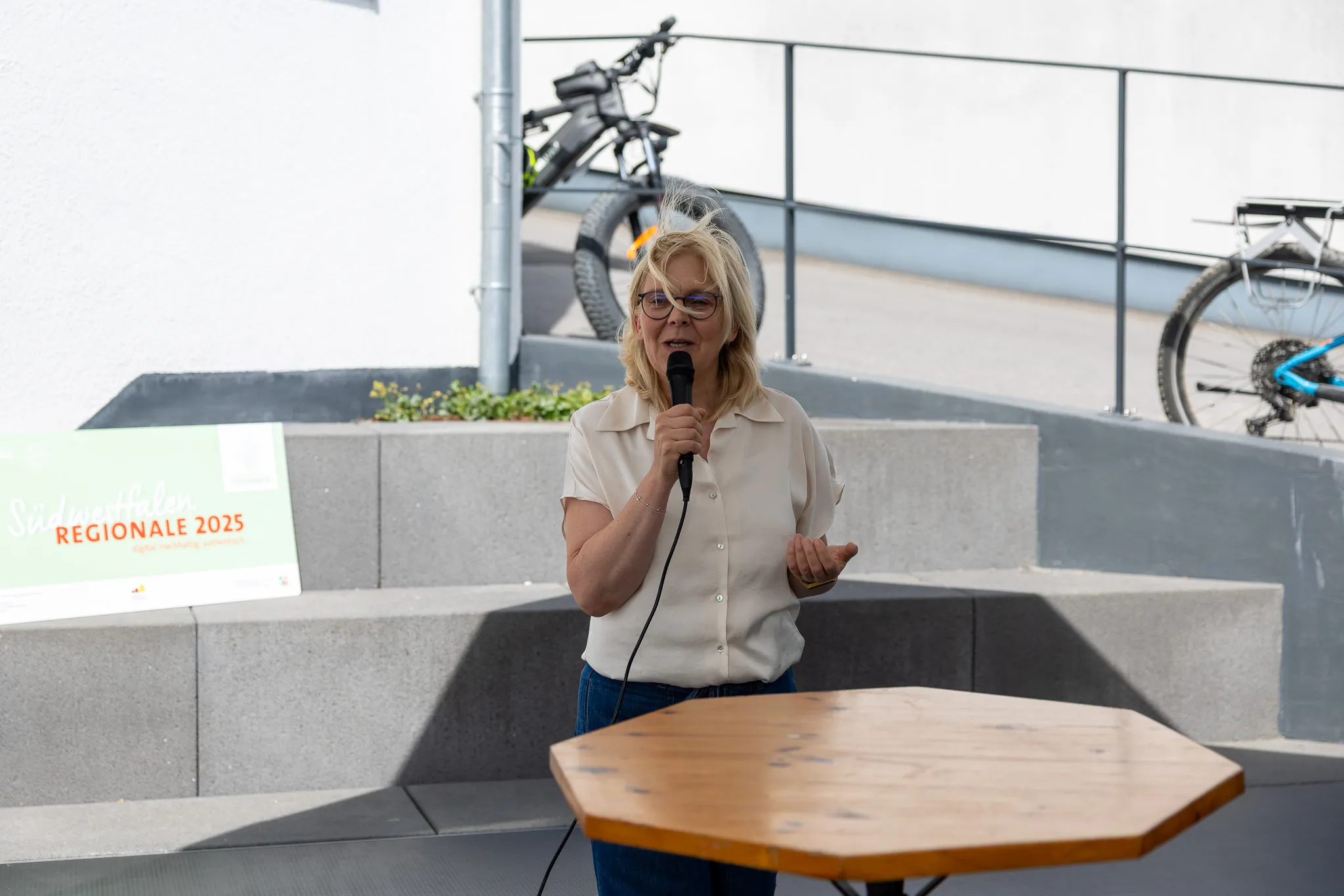 Eine Frau mit blonden Haaren hält ein Mikrofon und spricht auf einer Veranstaltung vor Publikum. Im Hintergrund sind Fahrräder und ein Banner mit der Aufschrift „Regionale 2025“ zu sehen.