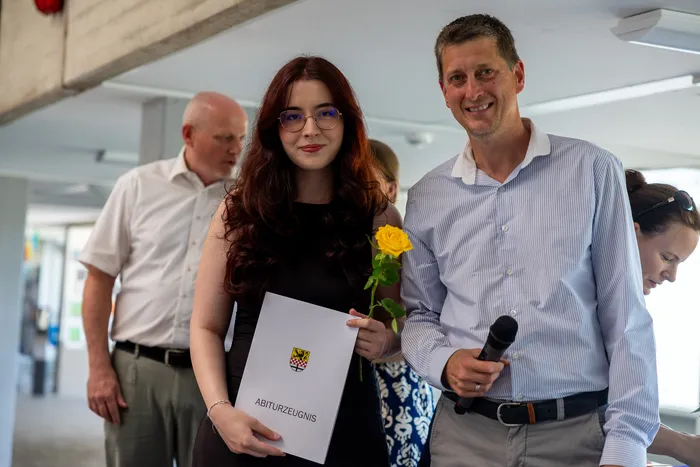 A young woman with long, curly red hair receives a high school diploma and a yellow rose from a smiling man holding a microphone. Two other people are visible in the background.