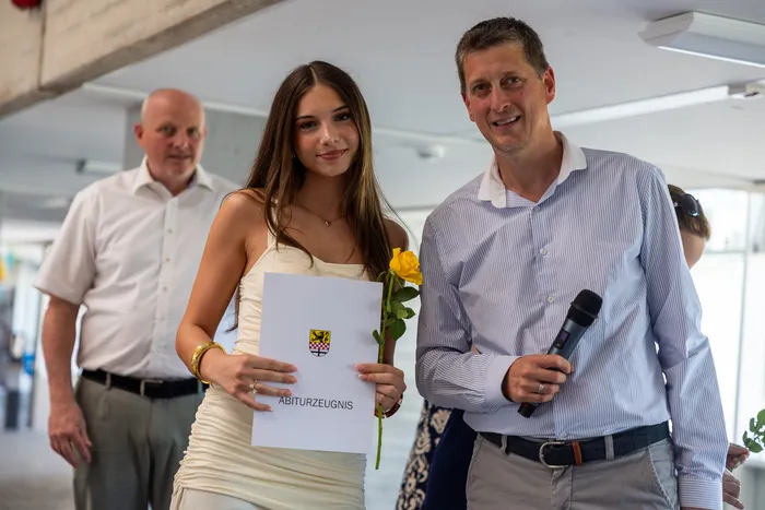 Eine junge Frau hält ihr Abiturzeugnis und eine gelbe Rose, während ein Mann neben ihr mit einem Mikrofon lächelt. Im Hintergrund steht ein weiterer Mann.