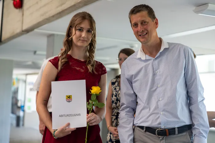 Eine junge Frau hält ihr Abiturzeugnis und eine gelbe Rose. Neben ihr steht ein Mann und lächelt in die Kamera.