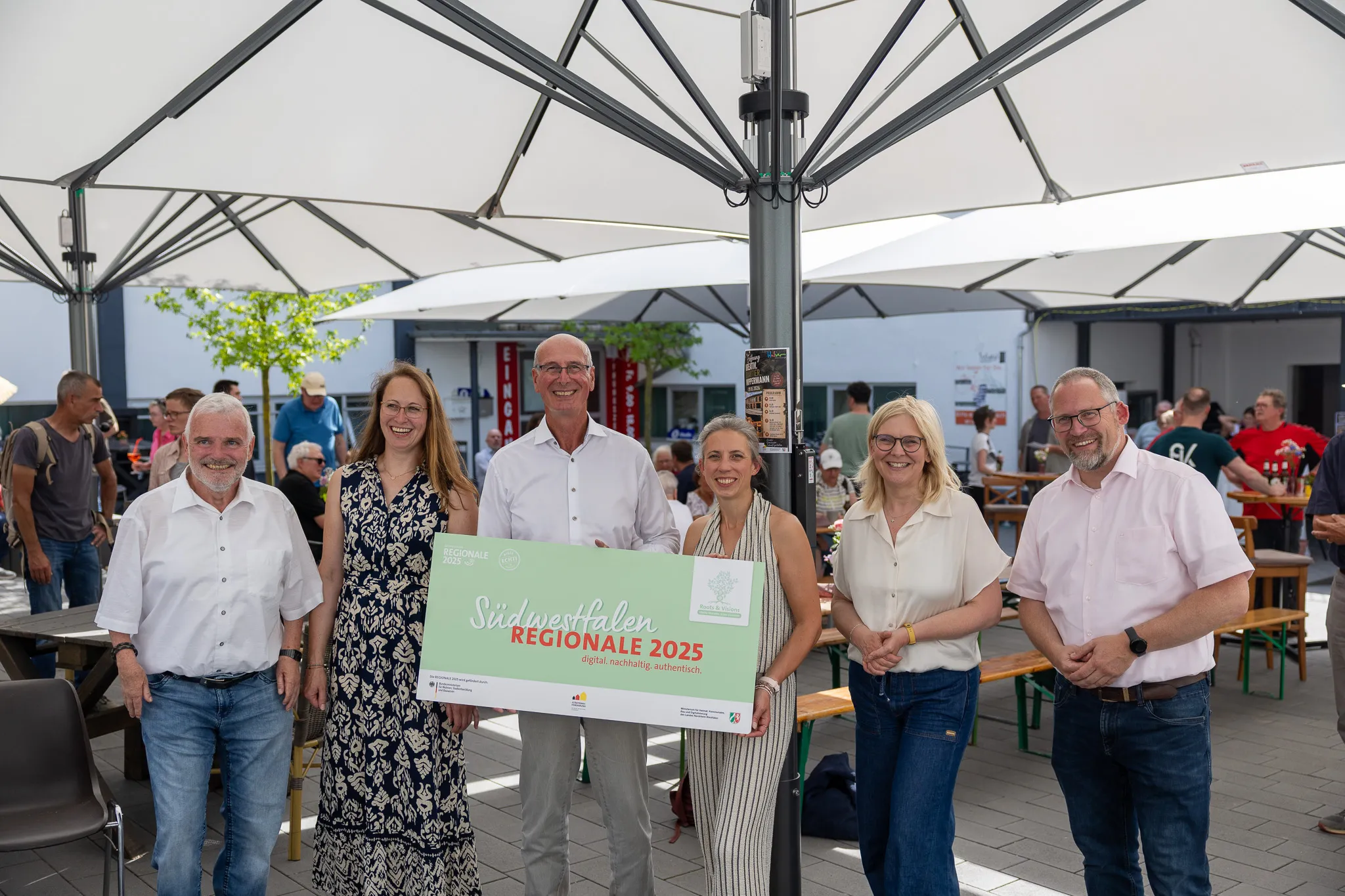 Sechs Personen stehen lächelnd vor einem Banner mit der Aufschrift „Südwestfalen REGIONALE 2025“ und dem Logo des Kreativ.Quartiers.Wippermann. Im Hintergrund sind weitere Menschen und ein Gebäude zu sehen.