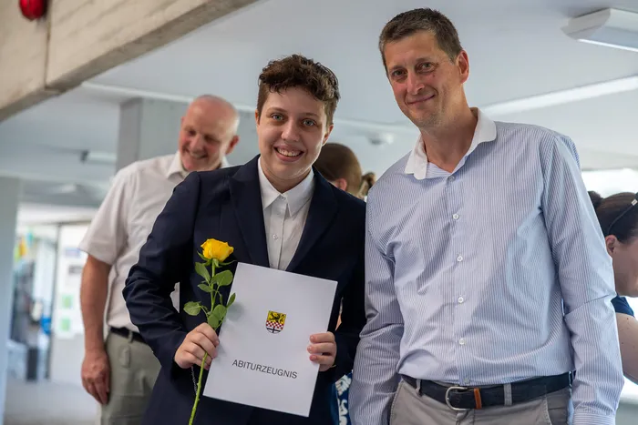 Ein junger Mensch in einem Anzug hält eine gelbe Rose und ein Abiturzeugnis mit einem Wappen. Neben ihm steht ein lächelnder Mann mit Hemd. Im Hintergrund sind weitere Personen zu sehen.