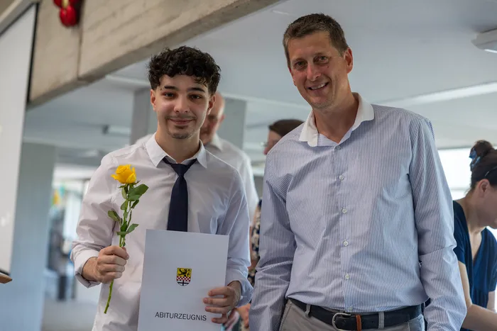 Ein junger Mann mit dunklen lockigen Haaren hält eine gelbe Rose und sein Abiturzeugnis mit dem Wappen von Nordrhein-Westfalen. Neben ihm steht ein lächelnder Mann mittleren Alters.