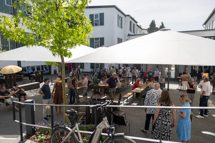Menschen stehen und sitzen auf einem gepflasterten Hof unter großen weißen Sonnenschirmen vor einem hellen Gebäude mit grünen Fensterläden. Im Vordergrund sind Fahrräder zu sehen.