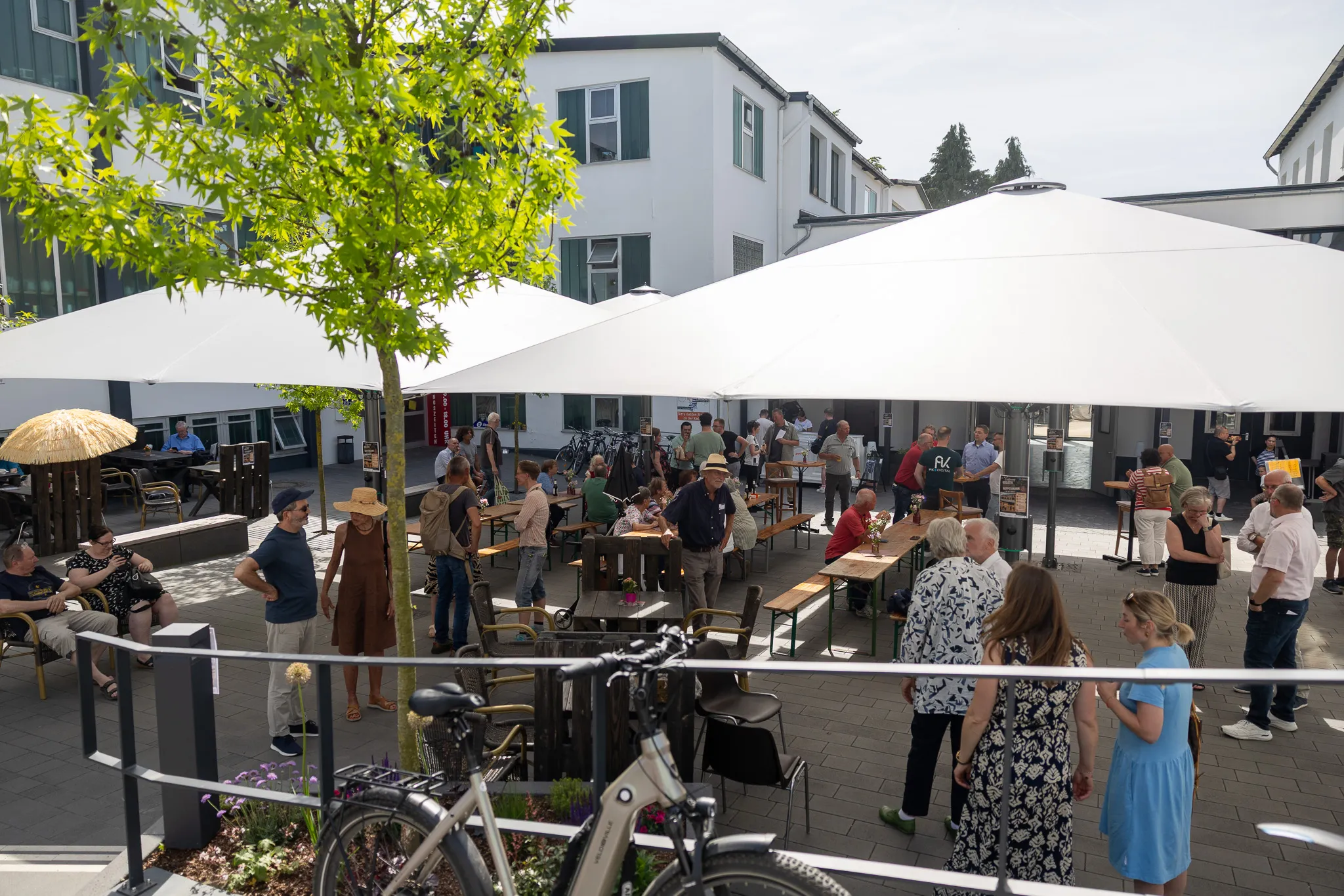 Menschen stehen und sitzen auf einem gepflasterten Hof unter großen weißen Sonnenschirmen vor einem hellen Gebäude mit grünen Fensterläden. Im Vordergrund sind Fahrräder zu sehen.