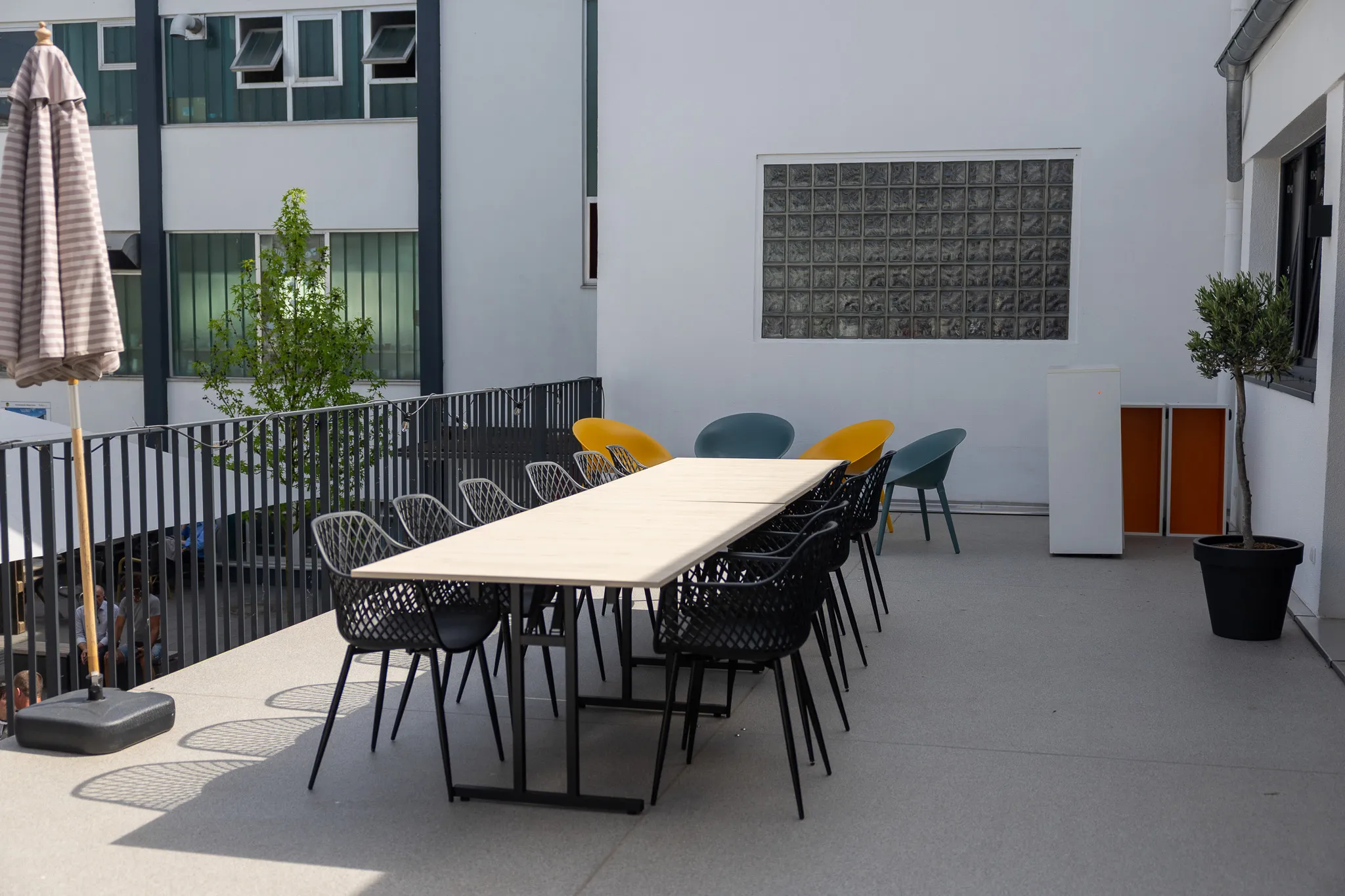 Ein langer Tisch mit mehreren Stühlen steht auf einer Dachterrasse vor einem modernen Gebäude. Im Hintergrund sind weitere Tische und eine Metallgeländer zu sehen.
