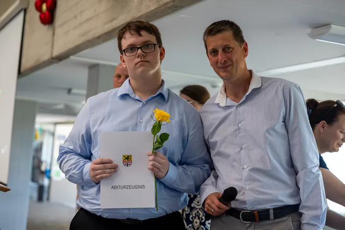 Ein junger Mann mit Brille hält ein Abiturzeugnis und eine gelbe Rose. Neben ihm steht ein lächelnder Mann mit Mikrofon.