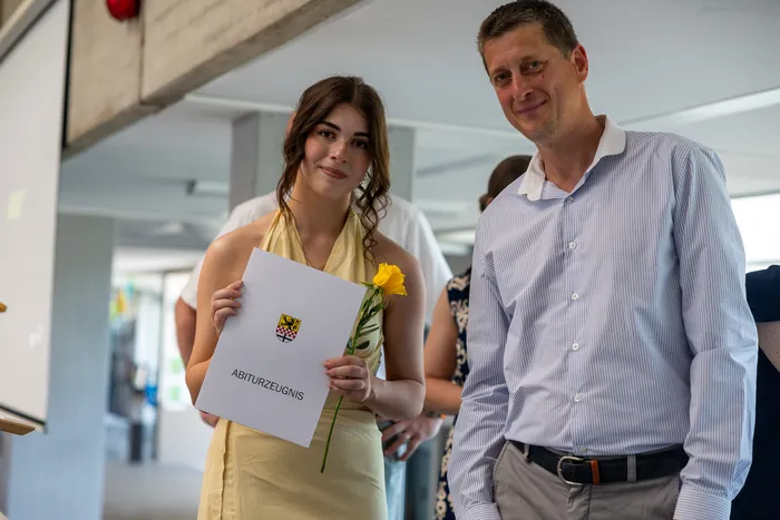 Junge Frau mit gelbem Kleid hält Abiturzeugnis und gelbe Blume. Neben ihr steht ein lächelnder Mann mit blau-weiß gestreiftem Hemd.