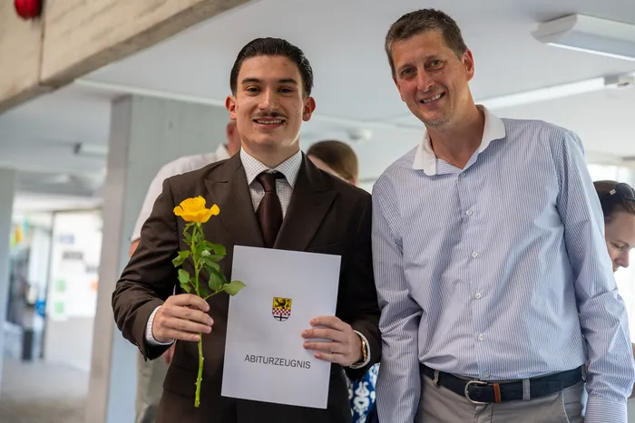 Ein junger Mann in Anzug und Krawatte hält eine gelbe Rose und sein Abiturzeugnis mit dem Wappen der Schule. Neben ihm steht ein lächelnder Mann in Hemd und Hose.