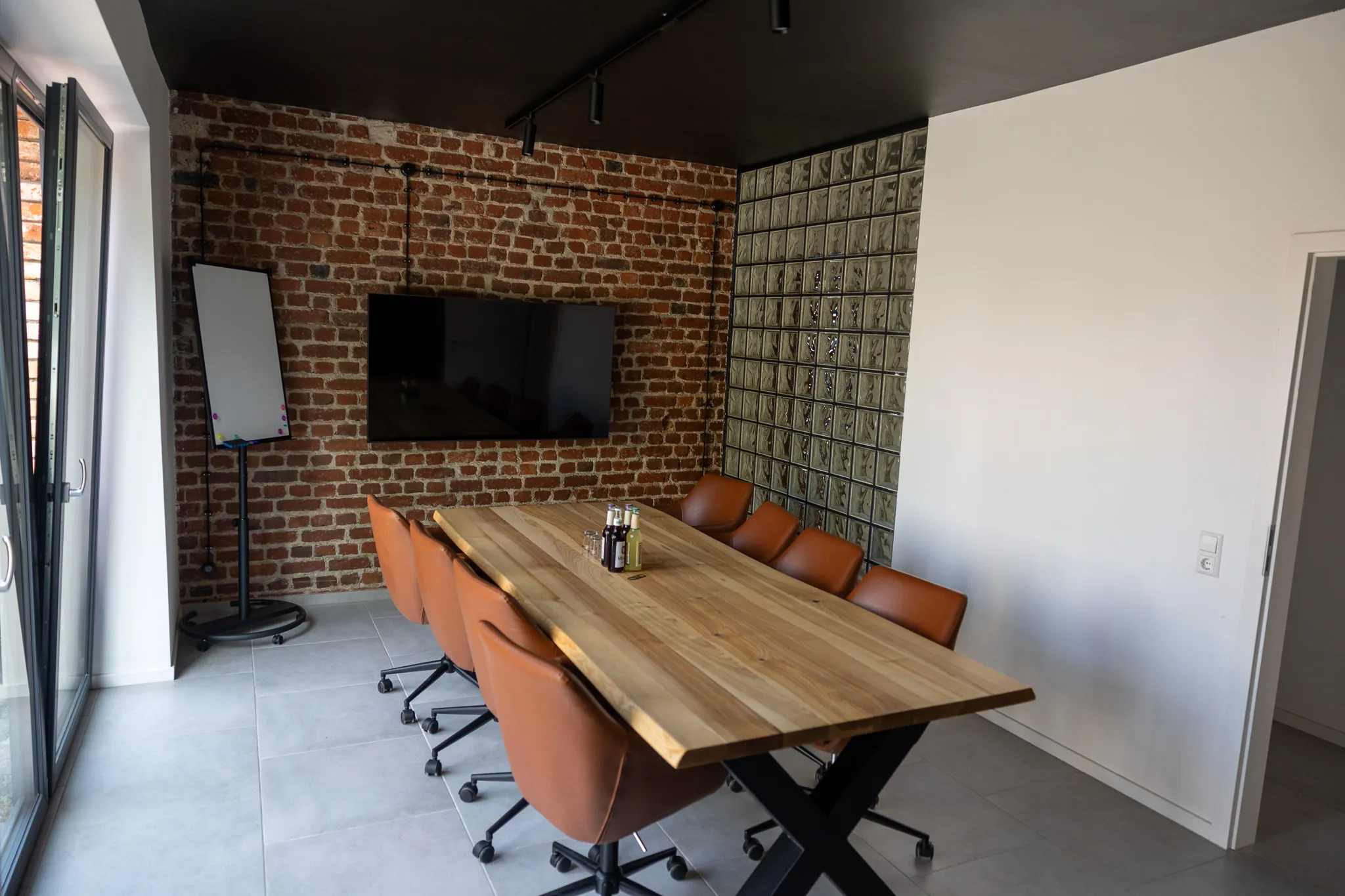 Ein heller Besprechungsraum mit einem großen Holztisch und braunen Ledersesseln. An der Wand ein Fernseher über einem Backsteinmauerwerk und eine Präsentationsfläche.