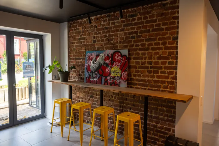 Ein Innenraum mit einer roten Backsteinwand, an der ein farbenfrohes Poster hängt. Davor stehen gelbe Barhocker vor einer Holztheke, links ein Fenster mit Glastür.