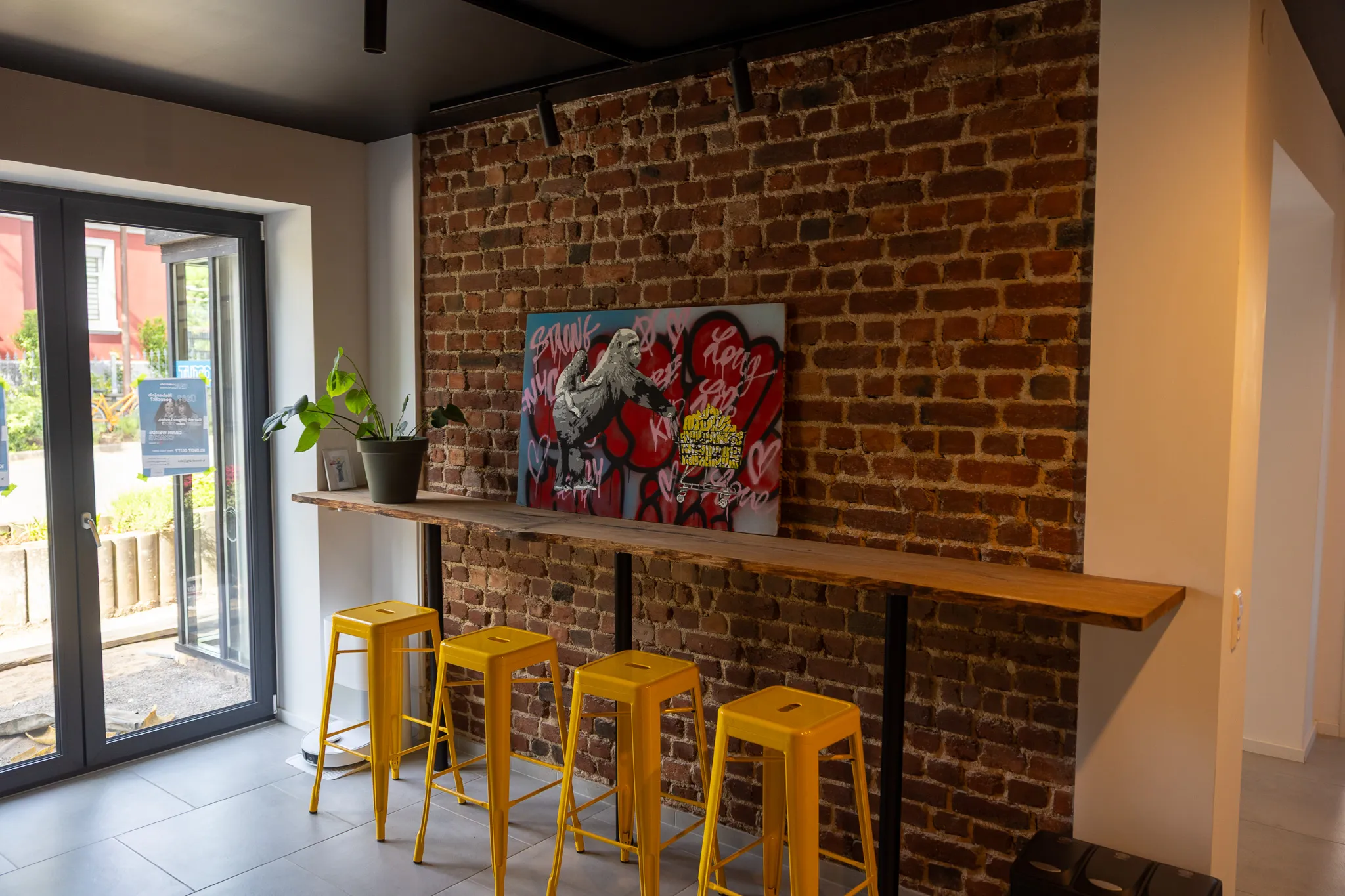 Ein Innenraum mit einer roten Backsteinwand, an der ein farbenfrohes Poster hängt. Davor stehen gelbe Barhocker vor einer Holztheke, links ein Fenster mit Glastür.