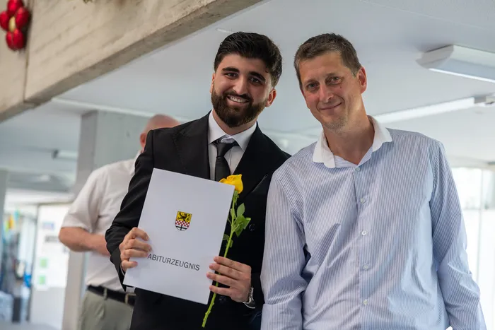 Ein junger Mann mit Bart hält eine Urkunde mit dem Aufdruck „Abiturzeugnis“ und eine gelbe Rose. Neben ihm steht ein lächelnder Mann in einem hellblauen Hemd.