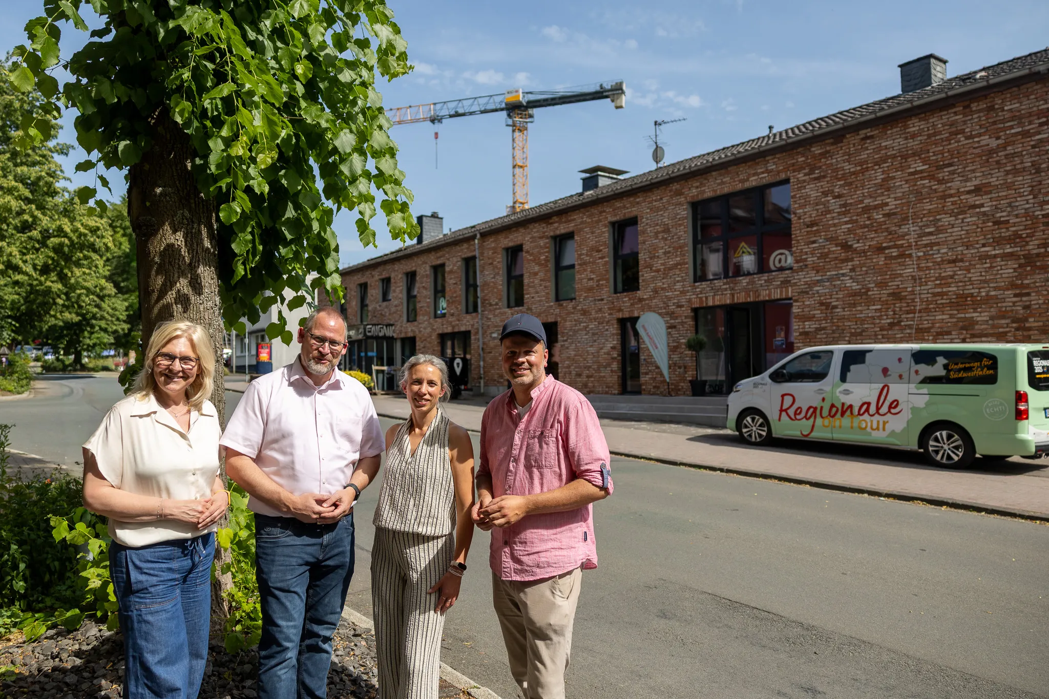 Vier Personen stehen vor einem Backsteingebäude mit Schaufenstern und einem Transporter mit der Aufschrift „Regionale Tour“. Im Hintergrund ein Kran.