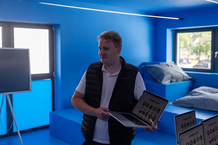 Ein Mann mit dunkler Weste hält einen Laptop und zwei Tablets mit ähnlichen Grafiken in der Hand, während er in einem hellblauen Raum steht.