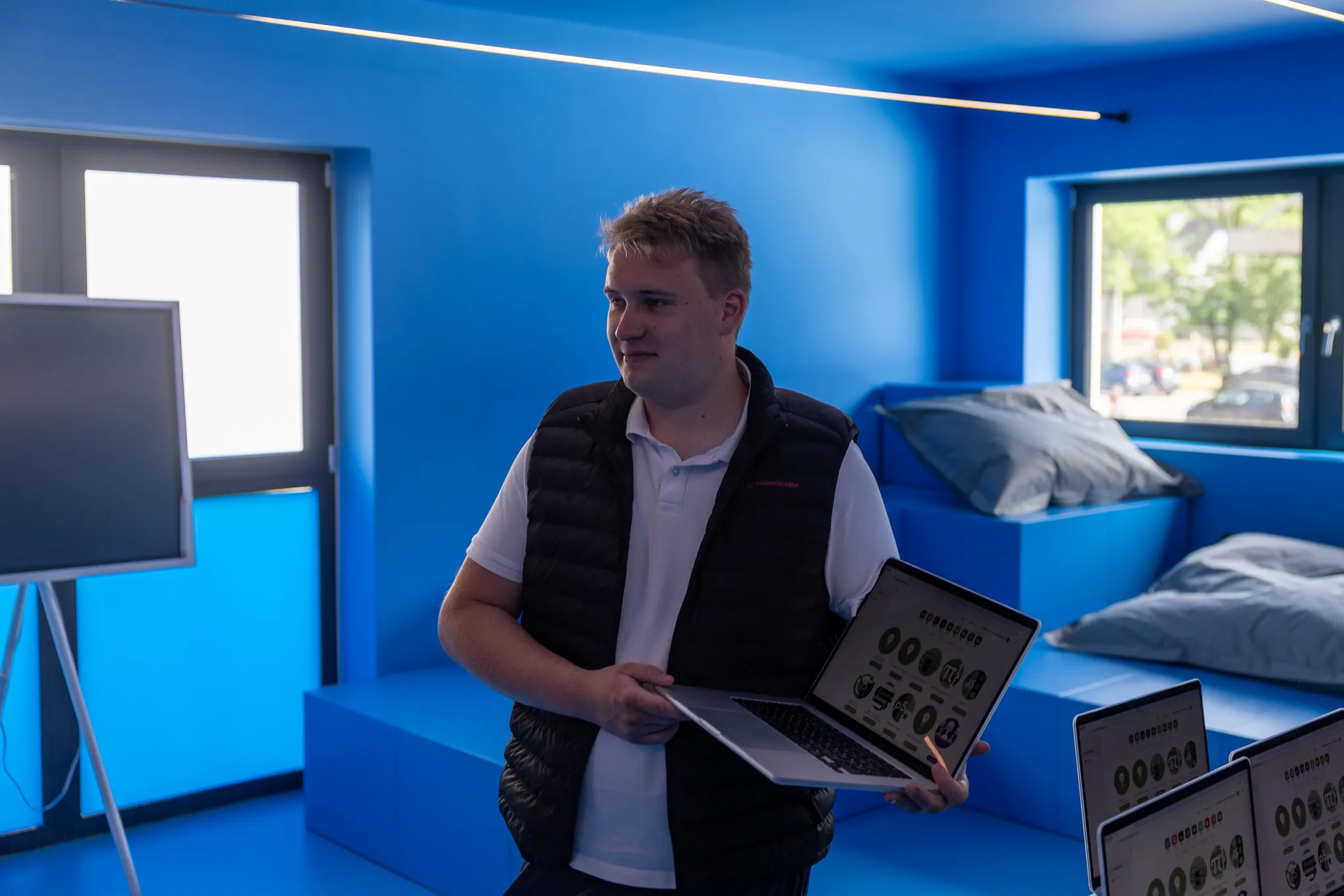 Ein Mann mit dunkler Weste hält einen Laptop und zwei Tablets mit ähnlichen Grafiken in der Hand, während er in einem hellblauen Raum steht.