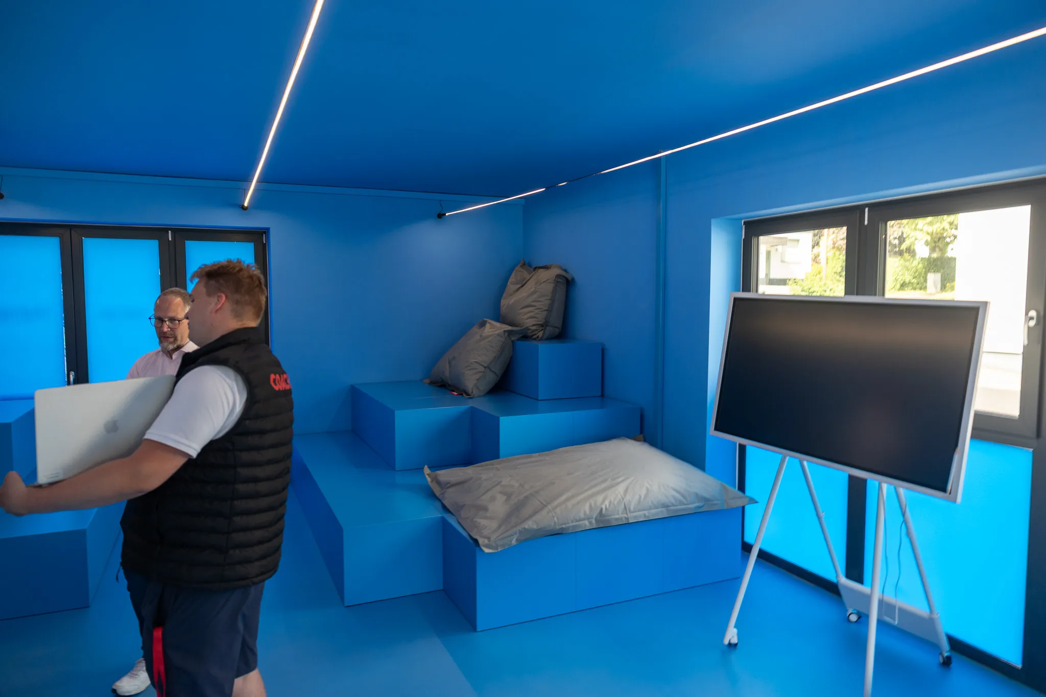 Ein Mann hält einen Laptop in einem hellblauen Raum mit mehreren blauen Würfeln als Sitzgelegenheiten. Im Hintergrund steht ein Mann und ein schwarzes Whiteboard.