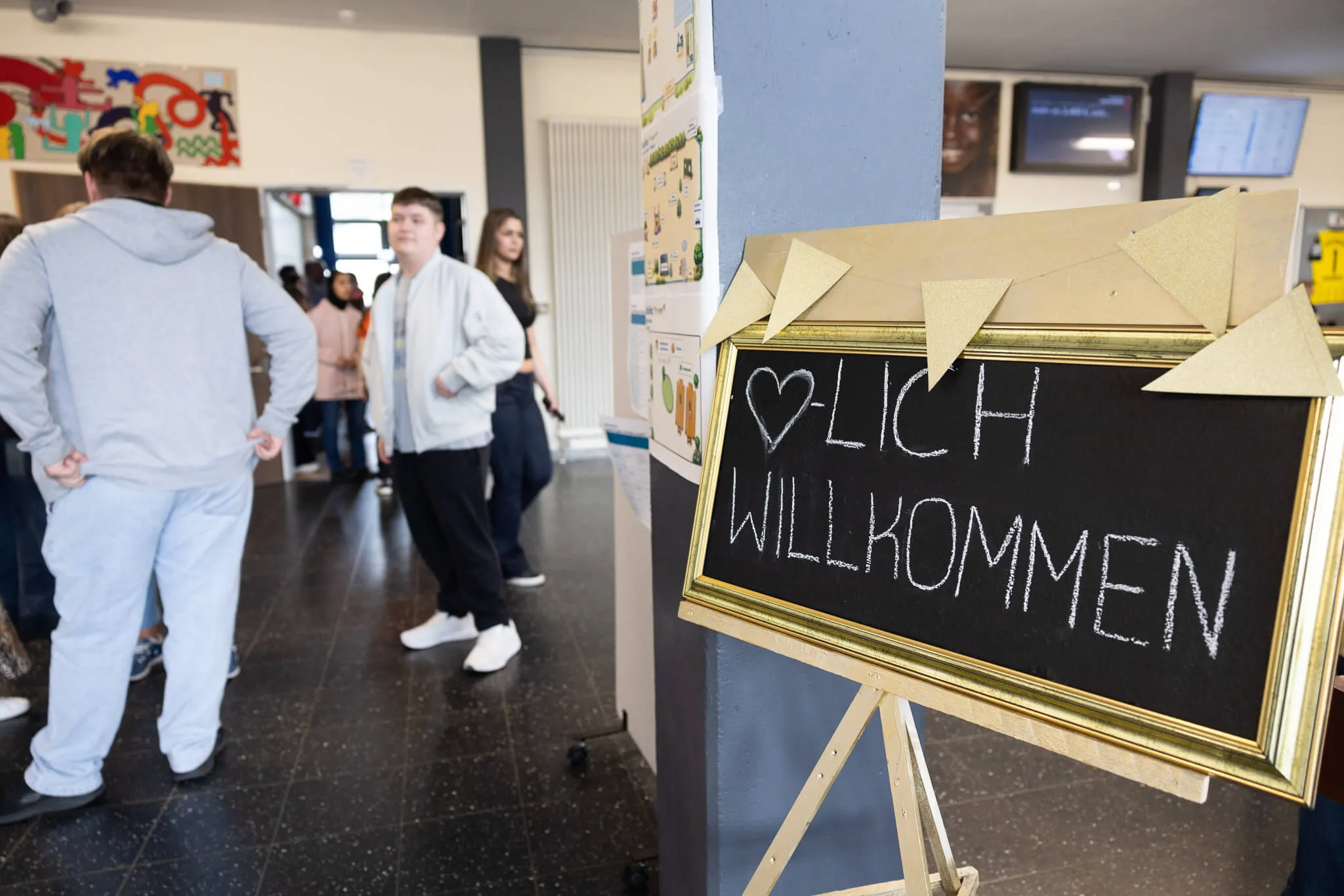 Schild mit der Aufschrift „Herzlich Willkommen“ steht im Vordergrund einer Schulfest-Szene mit Schülern im Hintergrund.