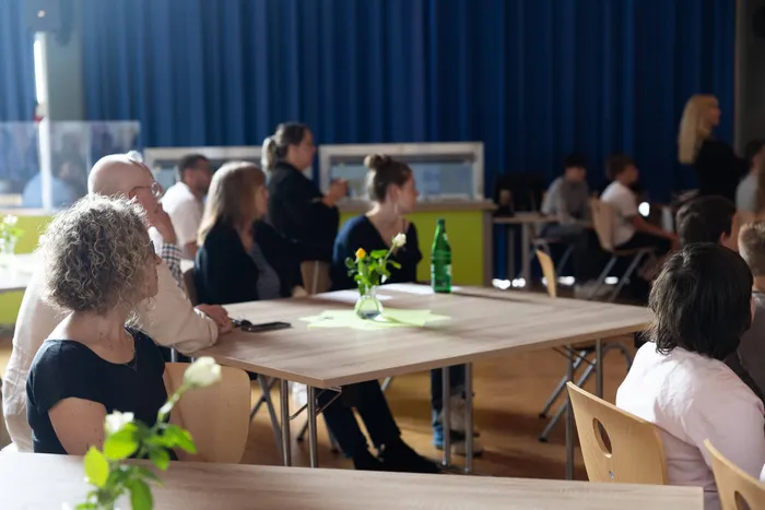 Menschen sitzen an Tischen in einer Turnhalle, einige unterhalten sich, andere hören zu. Im Hintergrund ein blauer Vorhang und weitere Personen.
