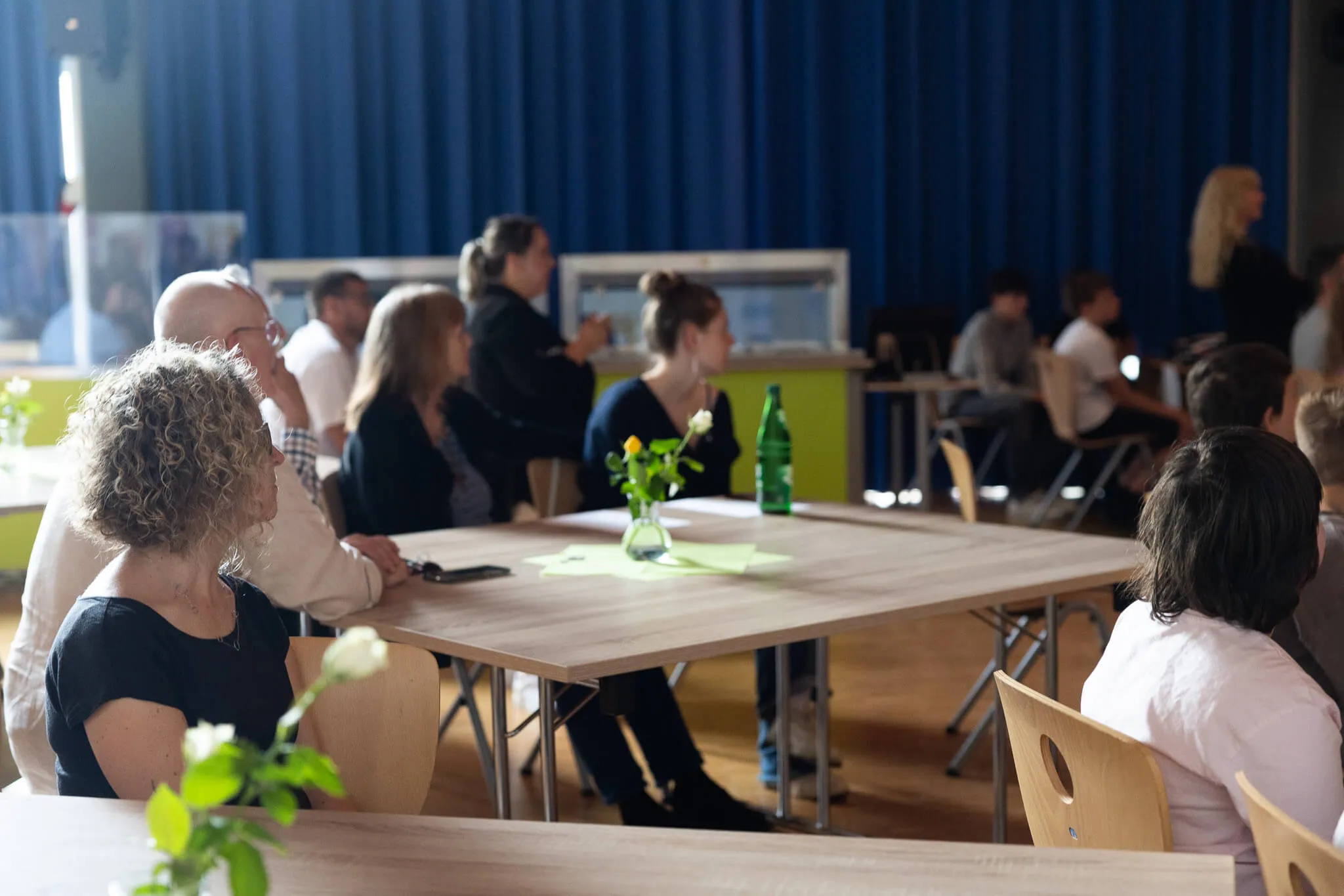Menschen sitzen an Tischen in einer Turnhalle, einige unterhalten sich, andere hören zu. Im Hintergrund ein blauer Vorhang und weitere Personen.