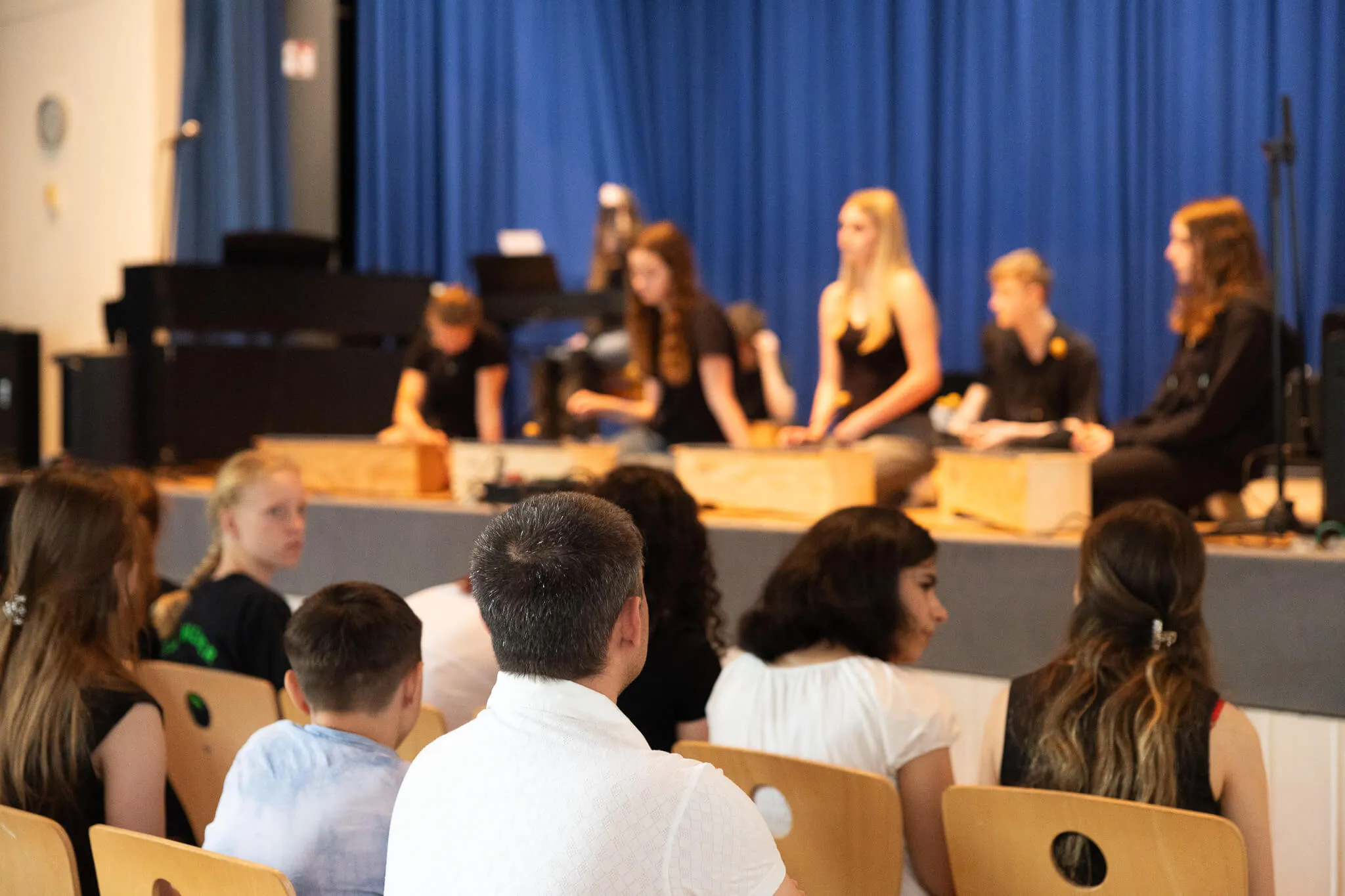 Schülerinnen und Schüler sitzen hinter Tischen auf einer Bühne und präsentieren etwas, während im Publikum andere Schüler zusehen.