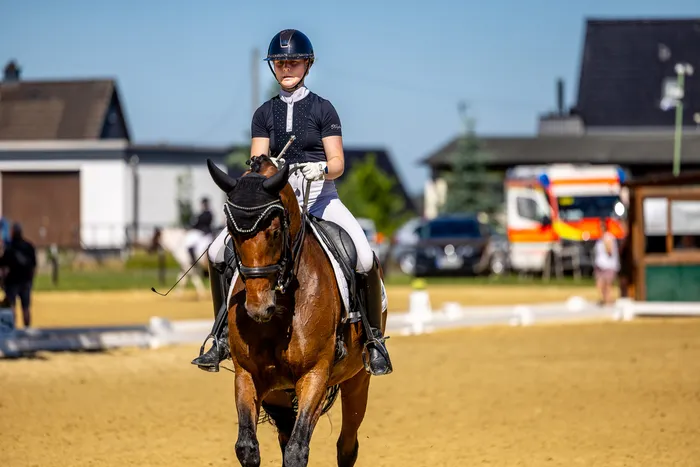 Eine Reiterin mit Helm und Jacke reitet auf einem braunen Pferd in einer Dressurprüfung. Im Hintergrund sind weitere Reiter und Gebäude zu sehen.