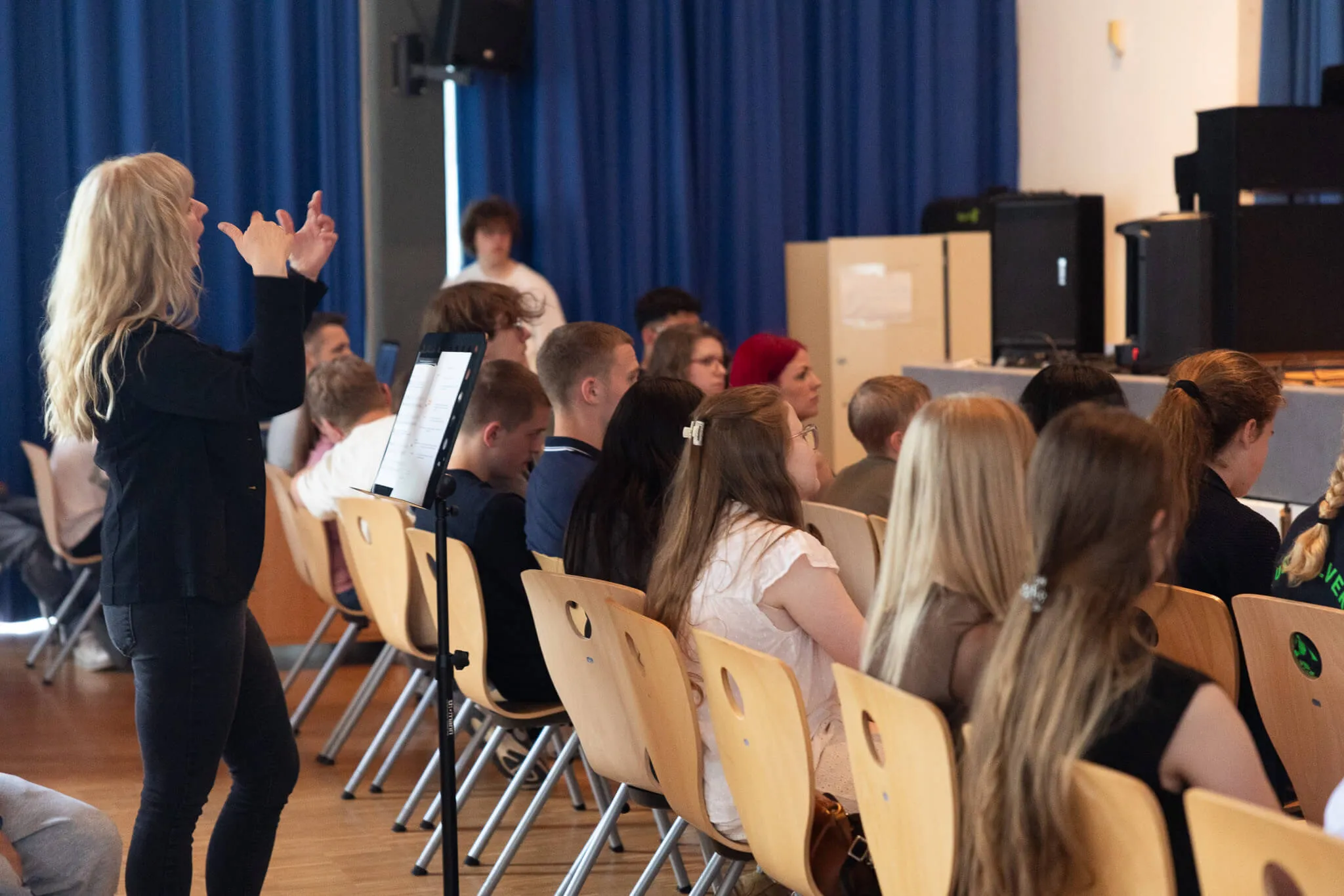 Eine blonde Frau dirigiert mit Handgesten vor einer Gruppe von Schülerinnen und Schülern in einem hellen Raum. Die Schüler sitzen auf Stühlen und verfolgen die Anweisungen.