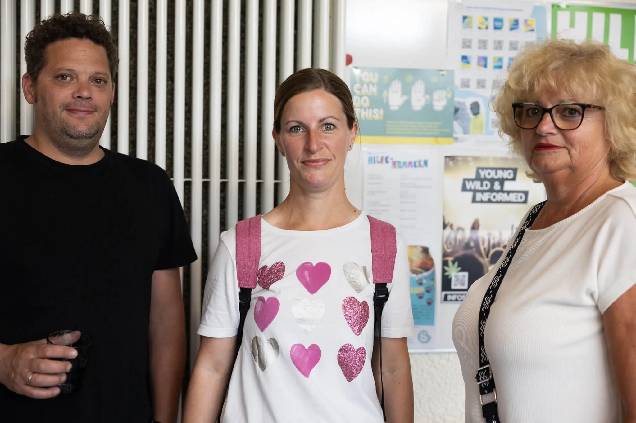 Drei Personen, ein Mann und zwei Frauen, stehen nebeneinander vor einem Plakat mit Informationen für Jugendliche. Im Hintergrund ist ein metallener Zaun zu sehen.