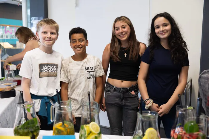Vier lächelnde Kinder stehen hinter einem Tisch mit bunten Getränken. Im Hintergrund sind weitere Personen und ein Schulfest zu sehen.